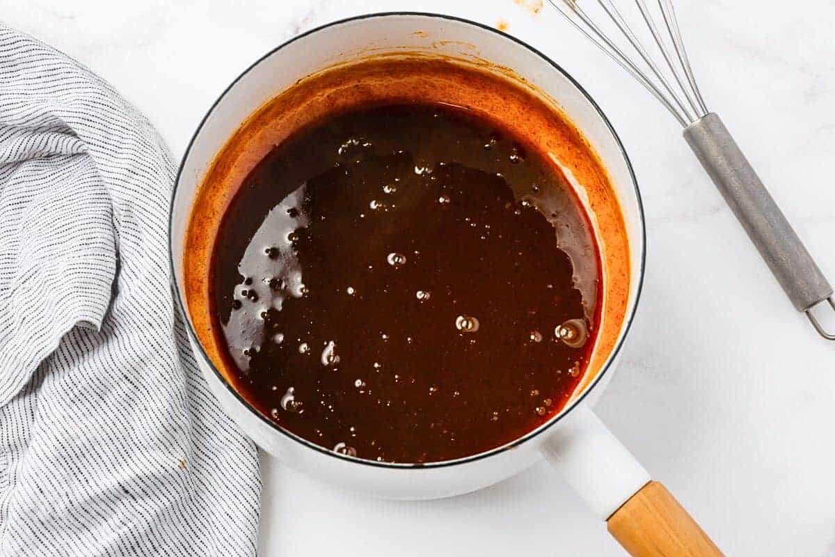 A white saucepan with a wooden handle contains a dark, bubbling sauce on a marble countertop. A striped kitchen towel and a whisk sit nearby.