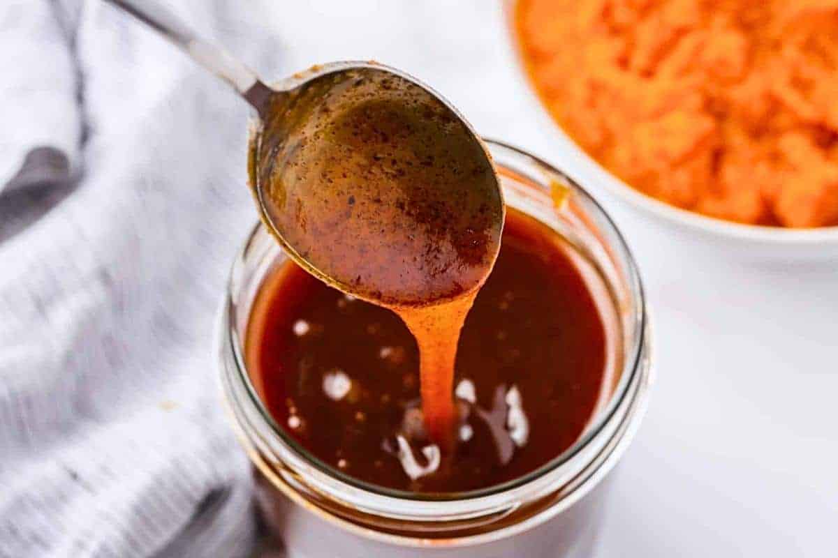 A spoon drips dark, thick sauce into a glass jar filled with the same sauce. In the background, there's a bowl with orange contents, possibly mashed sweet potatoes. A white cloth is partially visible on the side.