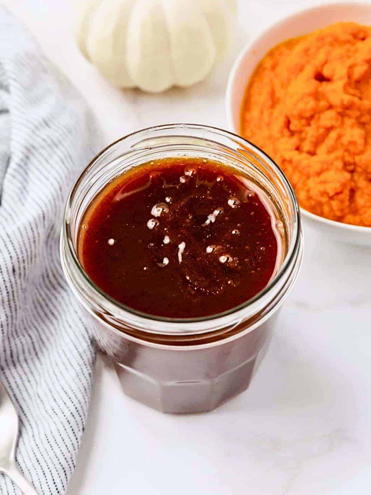 A glass jar filled with a dark amber sauce is placed on a white surface. Next to it, there's a blue-striped cloth and a white bowl containing orange puree. In the background, a white pumpkin is partially visible.