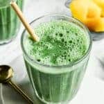 A glass of green smoothie with a straw on a white plate, next to a gold spoon. In the background, there are pieces of yellow fruit, likely mango, in a small dish. The smoothie appears frothy and vibrant.