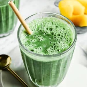 A glass of green smoothie with a straw on a white plate, next to a gold spoon. In the background, there are pieces of yellow fruit, likely mango, in a small dish. The smoothie appears frothy and vibrant.