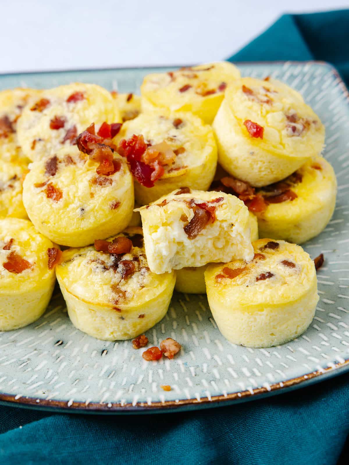 A plate of mini egg bites with bacon pieces on top. The eggs are fluffy and bite-sized, with one showing a bite taken out, revealing its soft interior. They are arranged on a textured square plate with a dark teal cloth underneath.