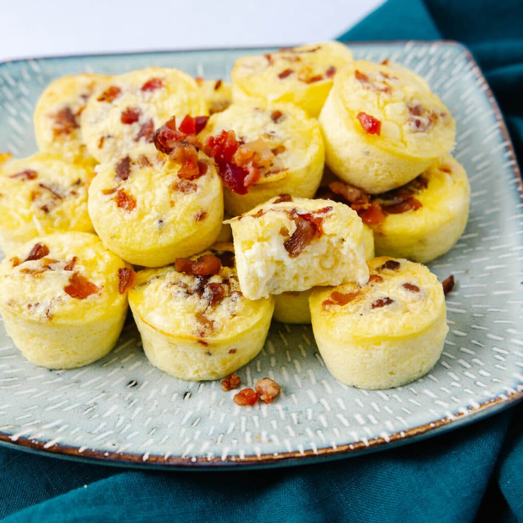 A plate of small egg bites topped with bits of bacon. The egg bites are arranged on a textured, light-colored plate resting on a dark green cloth. One egg bite is partially eaten, revealing its fluffy interior.