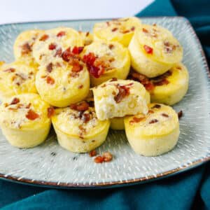 A plate of small egg bites topped with bits of bacon. The egg bites are arranged on a textured, light-colored plate resting on a dark green cloth. One egg bite is partially eaten, revealing its fluffy interior.