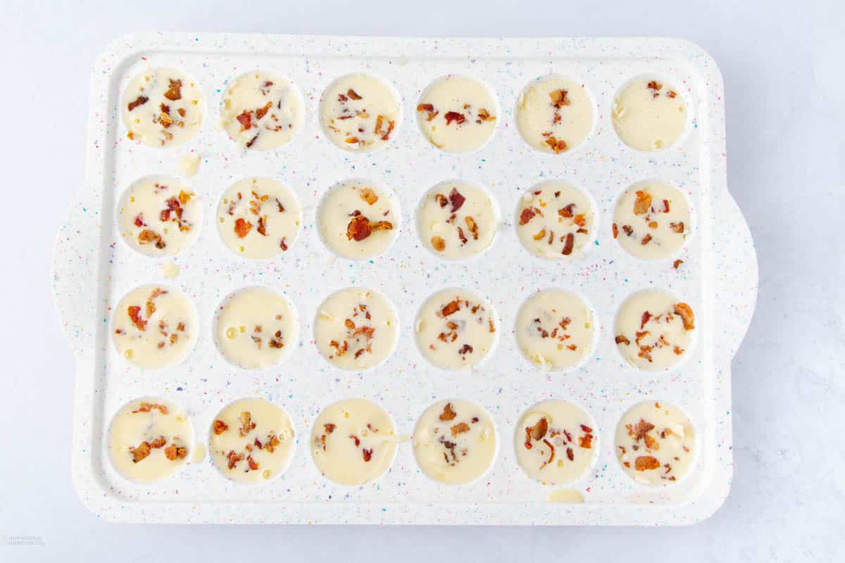 A white silicone mold with 24 round cavities filled with a creamy mixture topped with small, crumbled bacon pieces, ready to be frozen or baked.