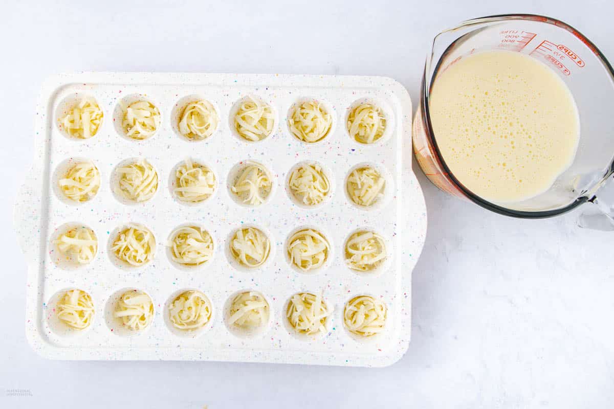 A white mini muffin tray filled with shredded cheese, next to a glass measuring cup containing a creamy mixture, placed on a light surface.