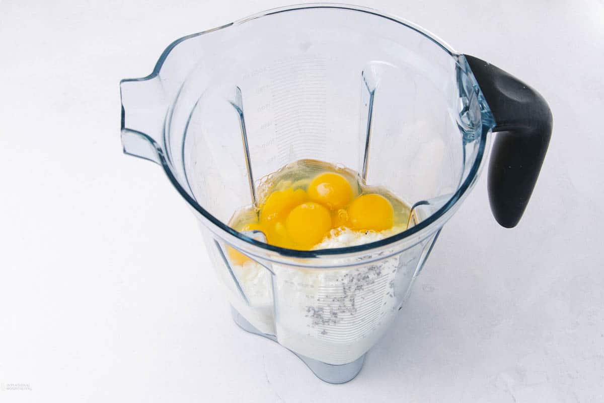 A blender pitcher containing raw eggs, flour, milk, and a pinch of seasoning, ready for blending.