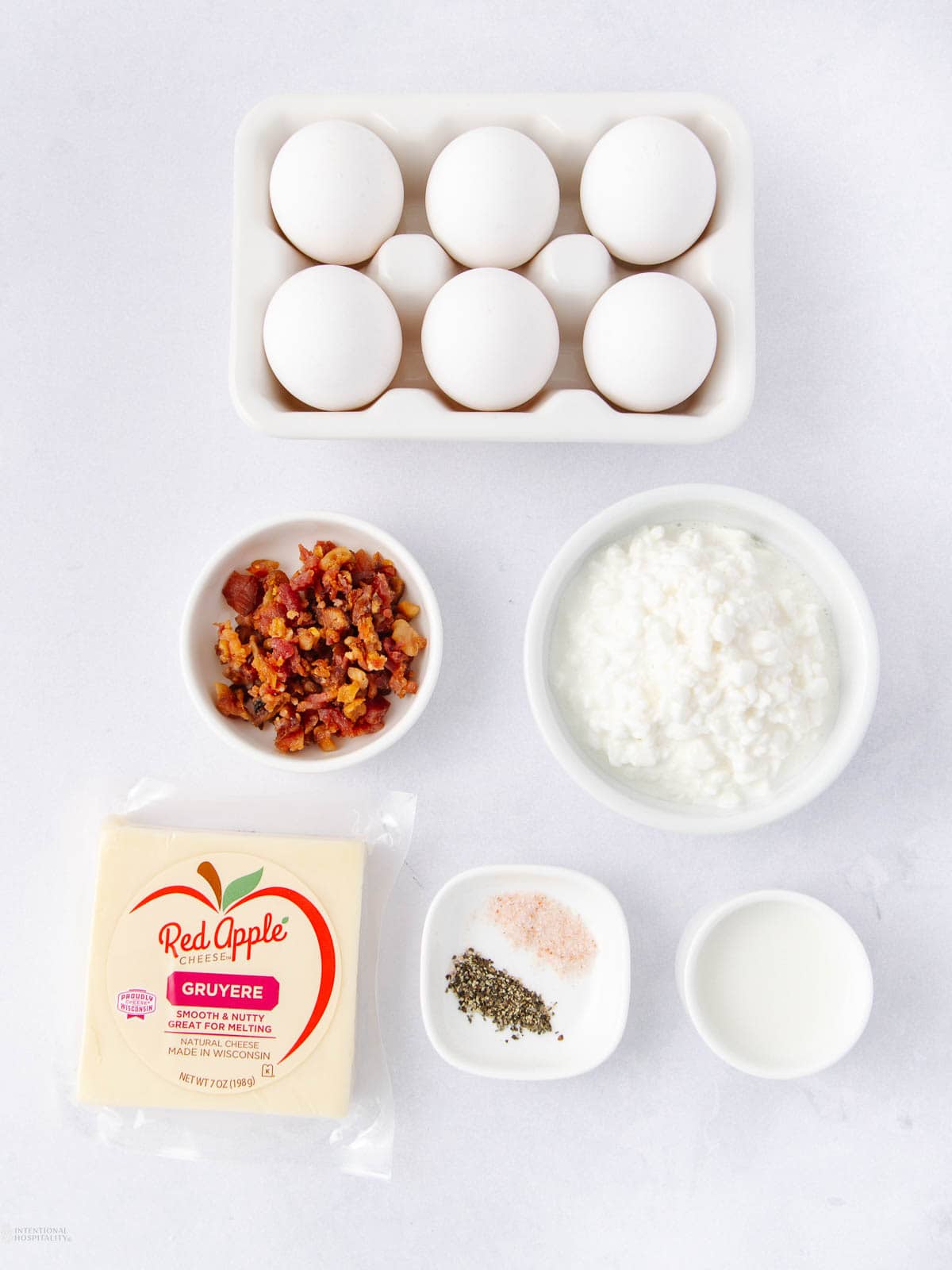 Top view of cooking ingredients on a white surface: a tray of six eggs, chopped bacon, cottage cheese, a block of Gruyere cheese, small bowls of pink salt, black pepper, and a splash of milk.