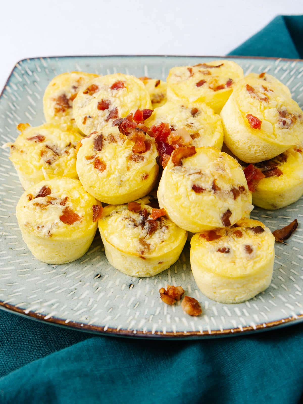 A plate of mini egg bites topped with crispy bacon pieces, placed on a textured plate. The plate is set on a teal cloth. The egg bites are golden and fluffy, arranged in a casual pile.