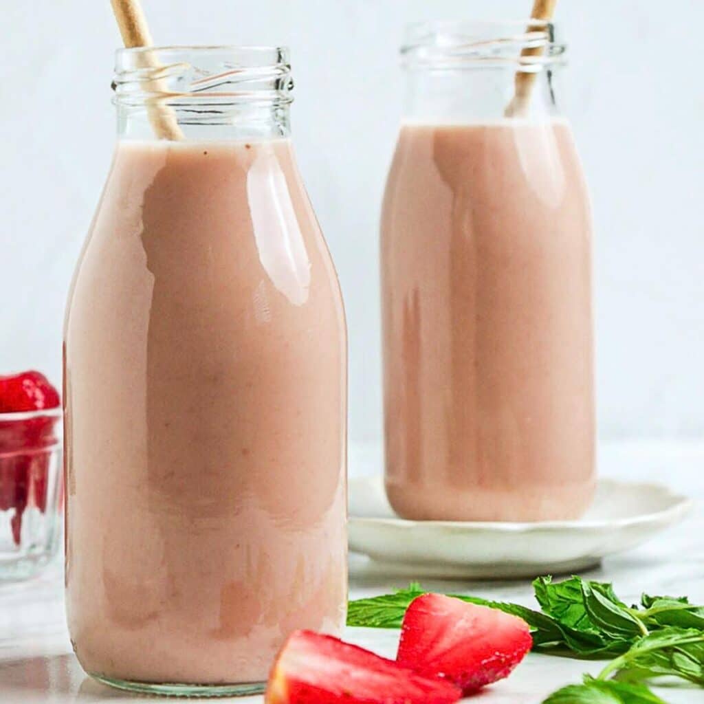 Two glass bottles filled with pink smoothies, each with a straw. They are placed on a white surface with fresh strawberries and mint leaves scattered around. A small glass bowl with strawberries is in the background.