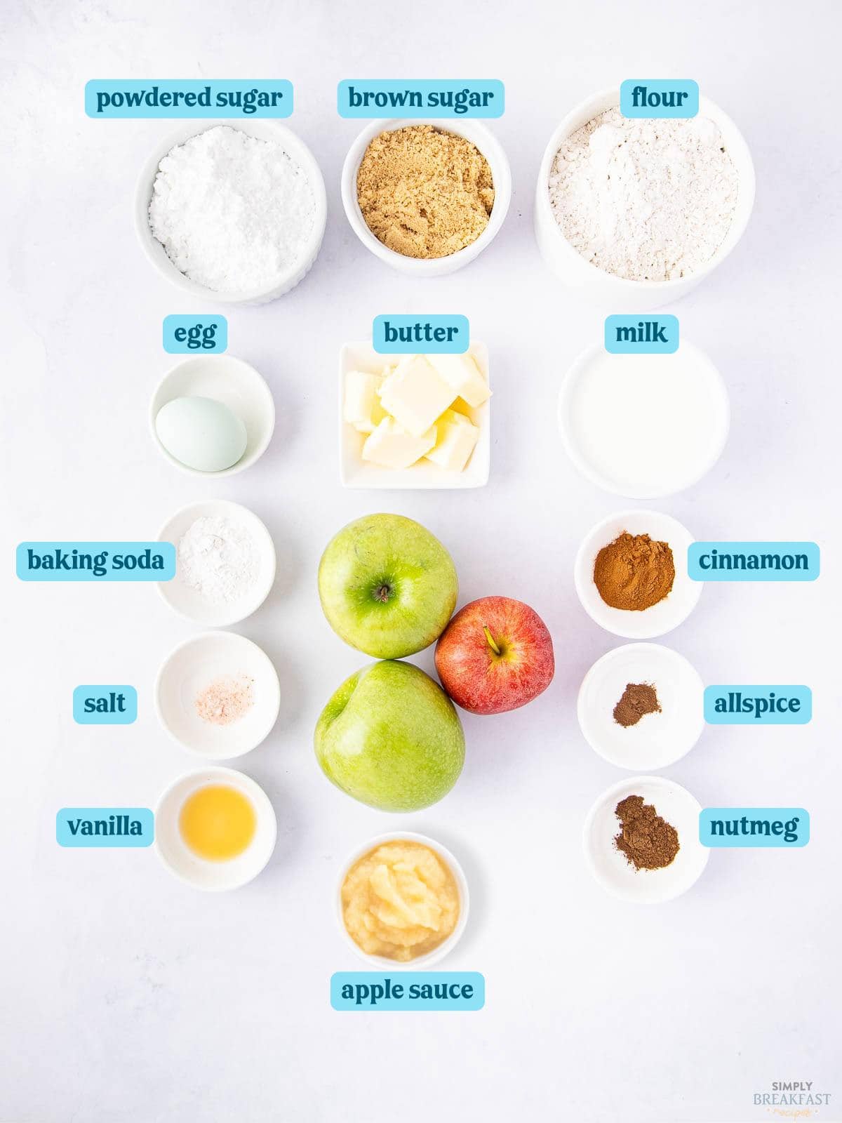 Top-down view of baking ingredients arranged on a light surface. Includes powdered sugar, brown sugar, flour, egg, butter, milk, baking soda, cinnamon, salt, allspice, vanilla, nutmeg, applesauce, and three apples (two green, one red).