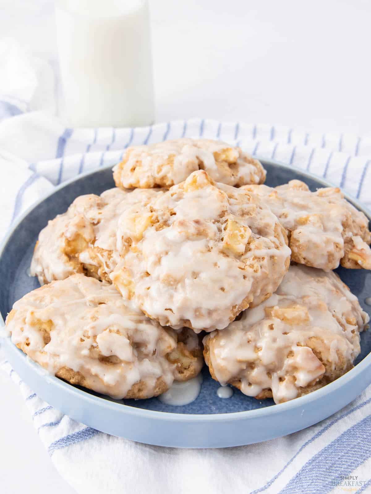 A blue plate filled with frosted apple cookies is shown. The cookies are glazed with a white icing and placed on a white and blue striped cloth. A glass of milk is visible in the background.