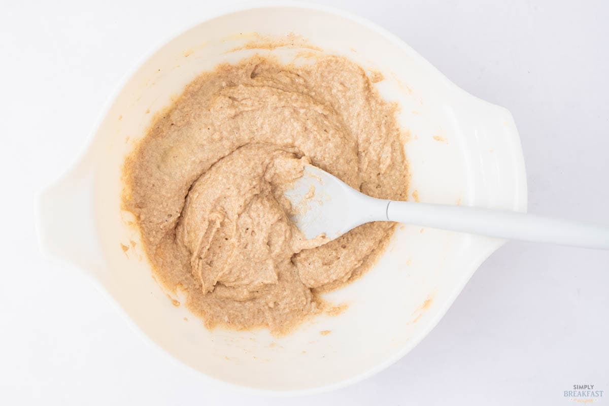 A white mixing bowl containing beige, thick batter with a white spatula resting inside. The batter appears partially stirred, with some streaks visible. The bowl is placed on a clean, white surface.