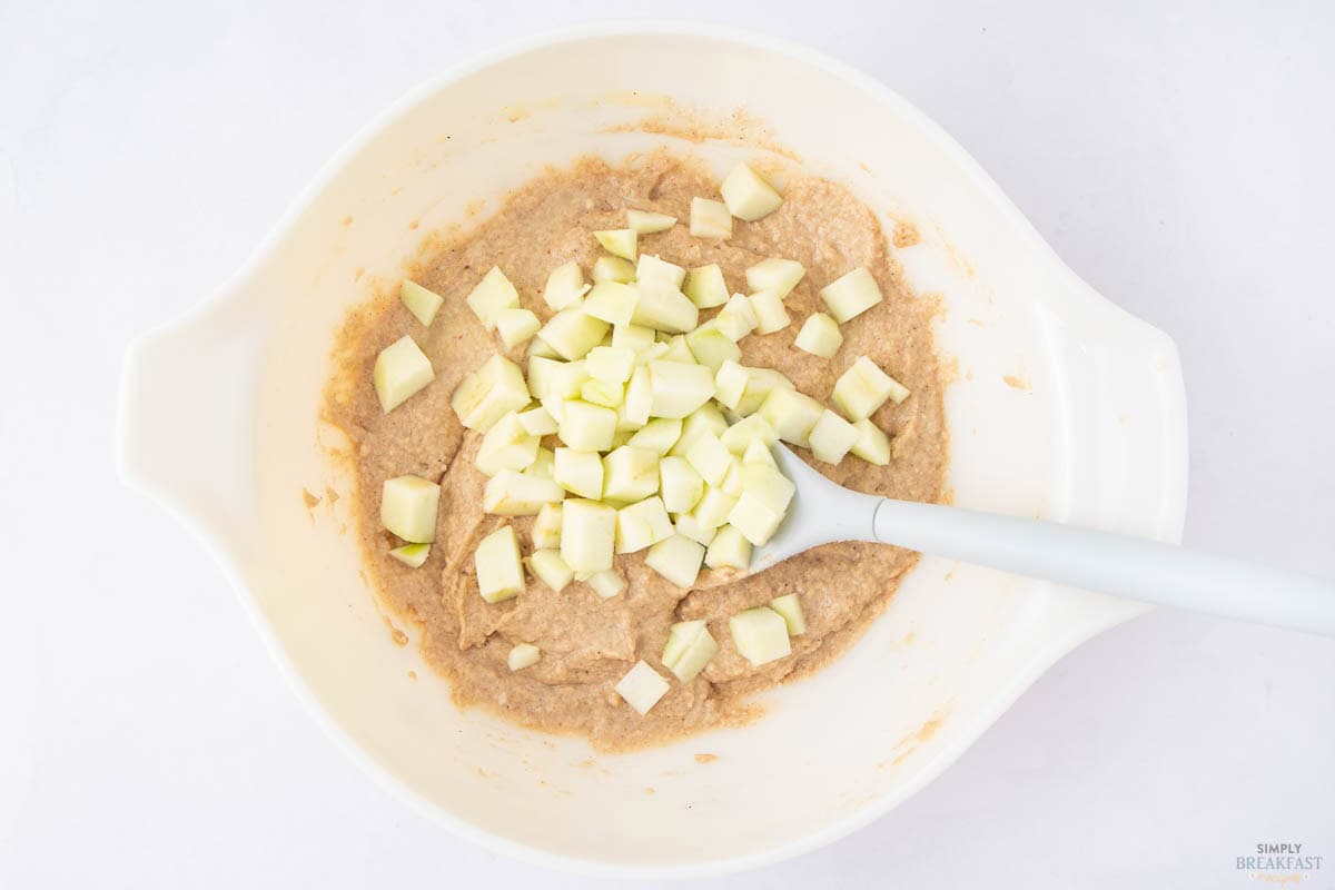 A white mixing bowl filled with brown batter and topped with chopped green apple pieces. A white spatula rests inside the bowl, which is placed on a white surface.