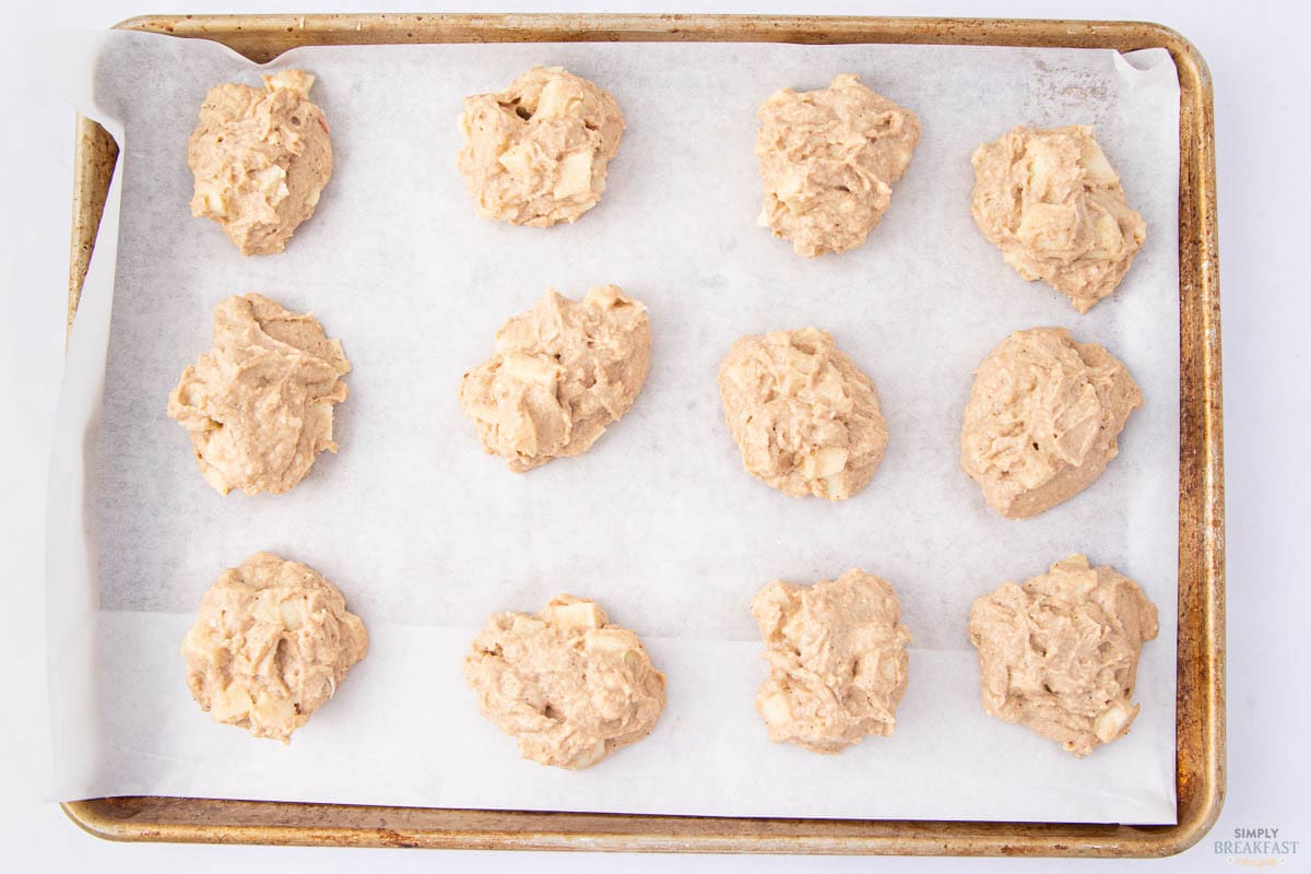 A baking sheet lined with parchment paper holds twelve dollops of uncooked cookie dough. The dough appears chunky, likely containing pieces of fruit or nuts, and is evenly spaced on the tray for baking.