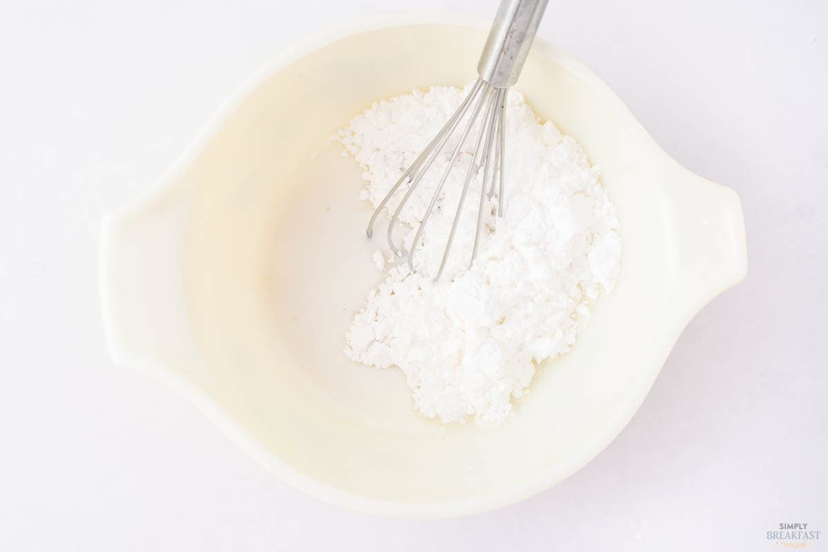 A mixing bowl containing a pile of white powder, likely flour or sugar, sits on a white surface. A metal whisk is partially submerged in the powder, ready for mixing.