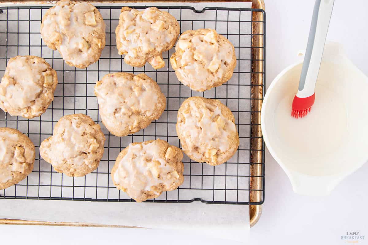 Icing-coated apple cookies cooling on a wire rack over a baking sheet. A bowl of icing with a red pastry brush is next to them.