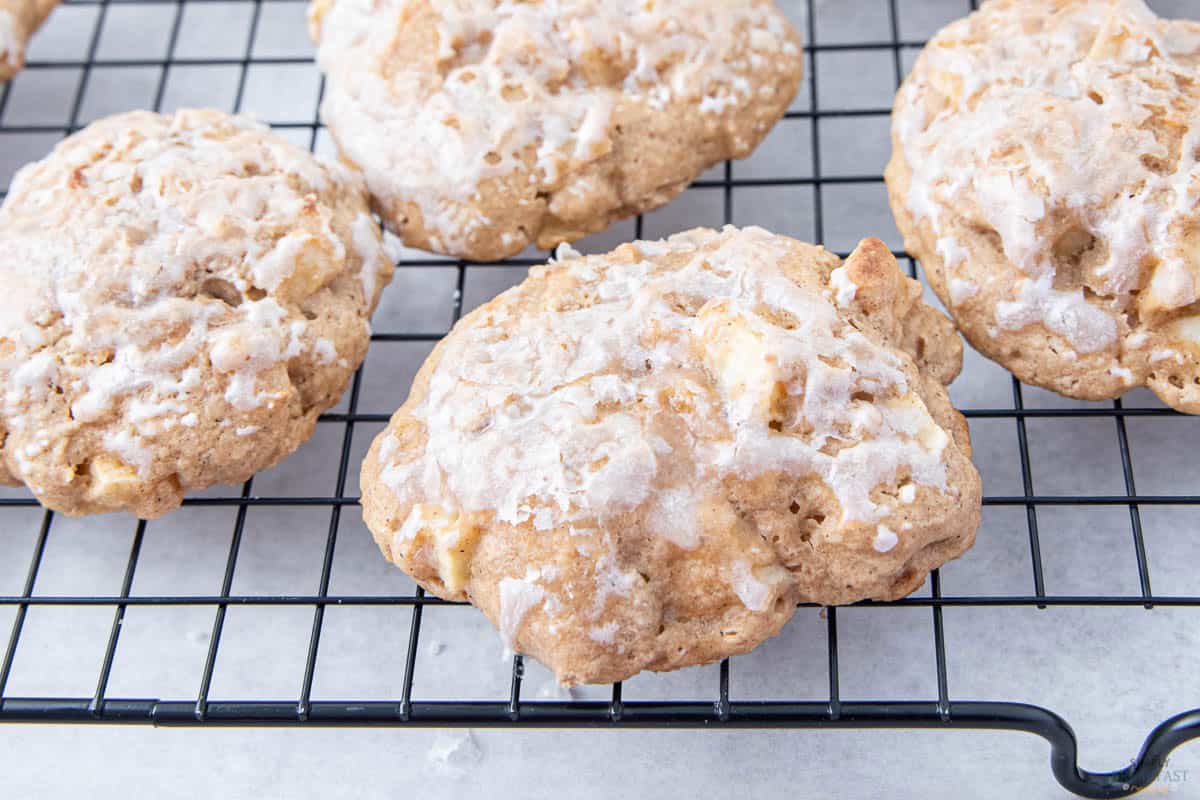 Close-up of apple cinnamon cookies with icing on a cooling rack. The cookies are golden brown with irregular shapes, and the icing adds a glossy texture on top, highlighting chunks of apple and a crumbly surface.