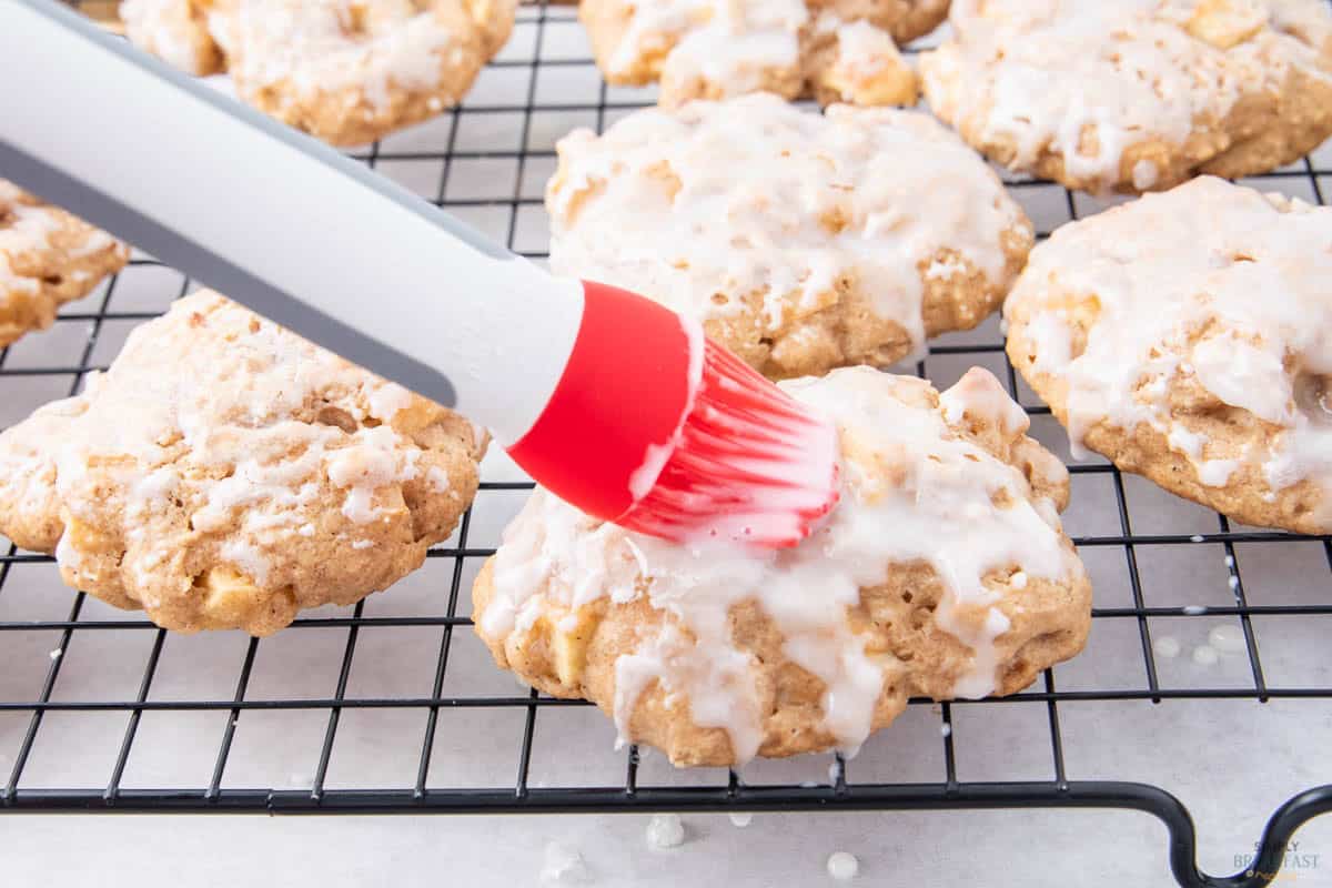 A red silicone brush is glazing icing onto oatmeal cookies with pieces of apple. The cookies are placed on a black wire cooling rack. The icing is white and glossy, covering most of the cookie surfaces.