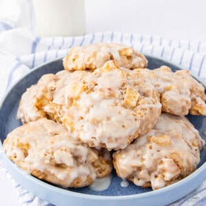 A blue plate holds a stack of glazed apple cookies with chunks of apple visible. A striped cloth and a glass of milk are in the background. The cookies have a shiny, sweet glaze covering them.