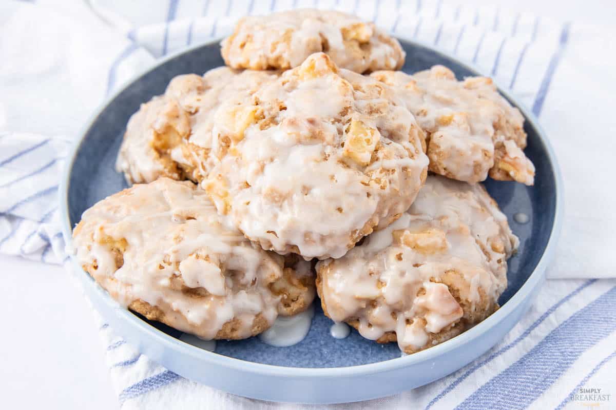 A blue plate with a stack of iced apple cookies. The cookies are glazed, showcasing chunks of apple and a light brown color. The plate is set on a white and blue striped cloth.