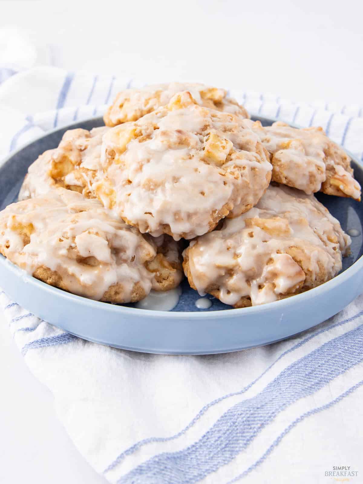 A blue plate filled with glazed apple fritters rests on a white and blue striped cloth. The fritters are coated in a light glaze, capturing a glossy sheen under the light.