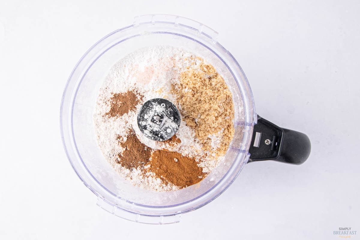 Top view of a food processor bowl filled with baking ingredients, including flour, brown sugar, and various spices like cinnamon and nutmeg, on a light surface.