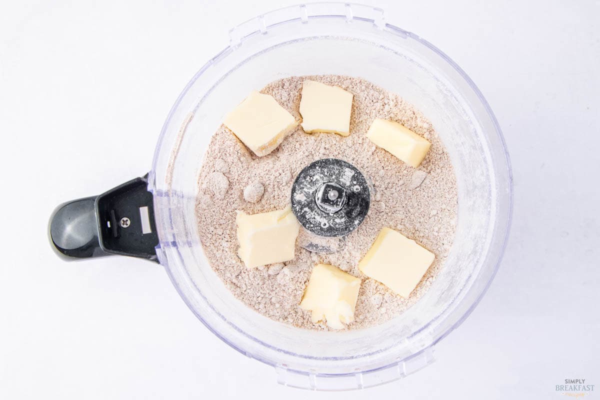 A food processor bowl containing flour and several cubes of butter, viewed from above. The processor blade is visible at the center. The background is light, enhancing the visibility of the ingredients.