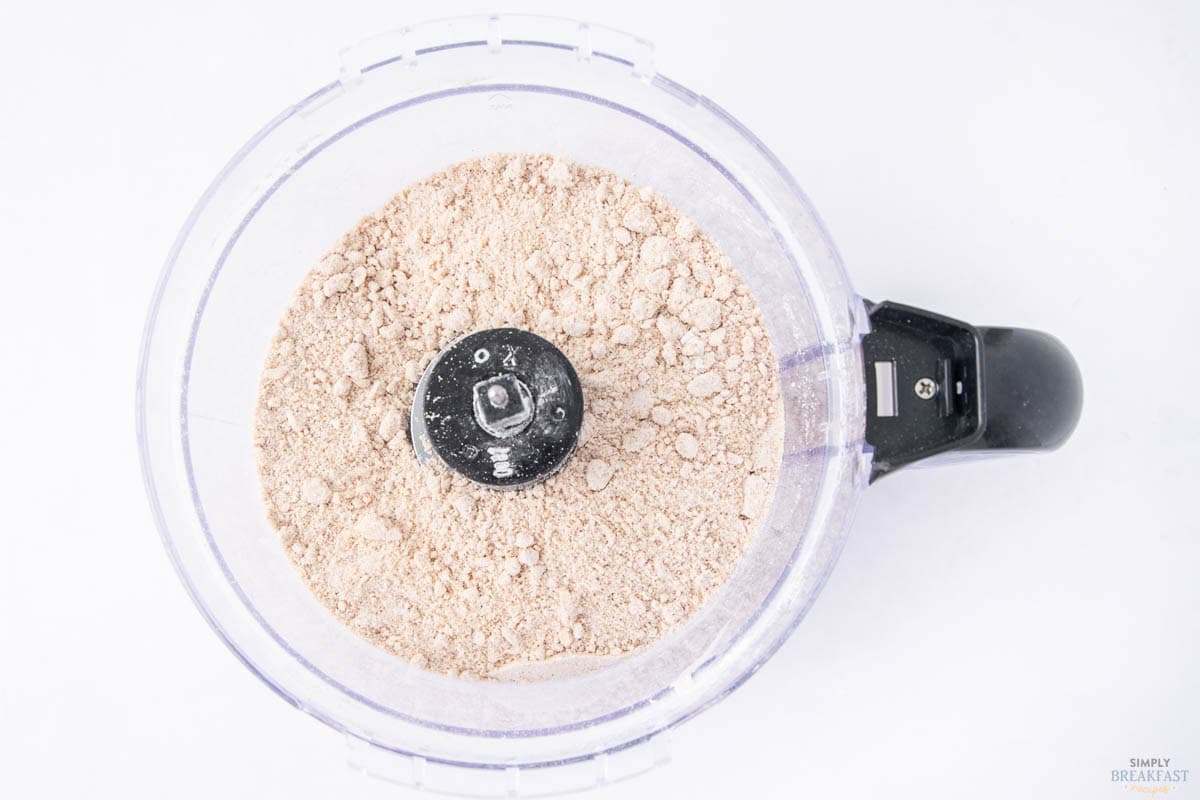 A clear food processor bowl containing a light brown, powdery mixture. The processor blade is visible at the center. The bowl rests on a white surface.