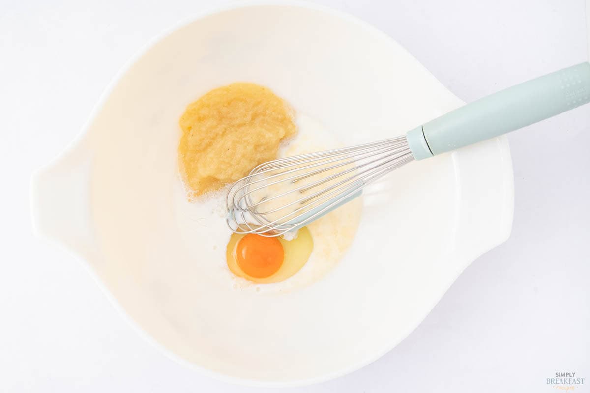 A white mixing bowl contains an egg yolk, applesauce, and other wet ingredients. A whisk with a light blue handle rests inside the bowl, positioned to mix the contents.