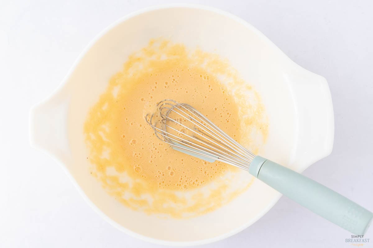 A white mixing bowl with a light orange batter being whisked. A metal whisk with a light blue handle rests inside. The batter shows signs of being mixed, with small bubbles visible on the surface.