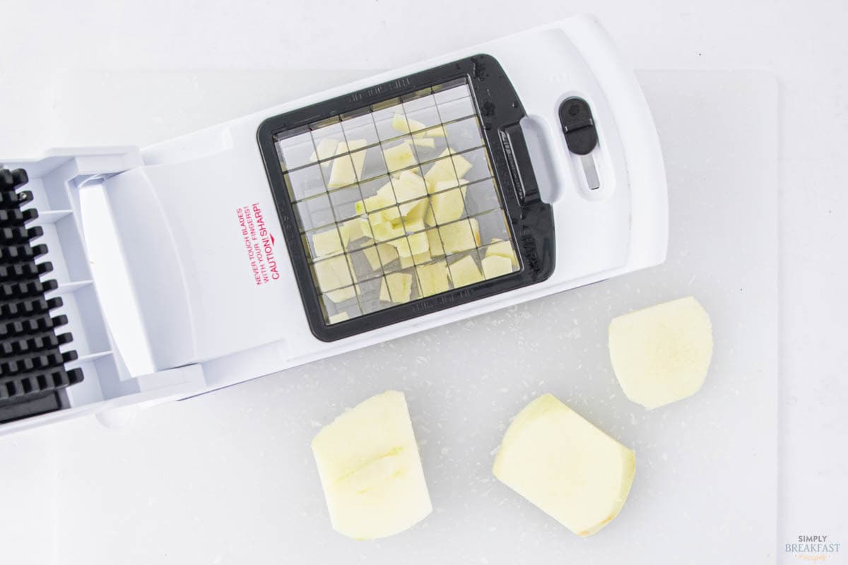 A white vegetable slicer with diced potatoes inside is placed on a white cutting board. Three larger pieces of potato are on the board next to the slicer.