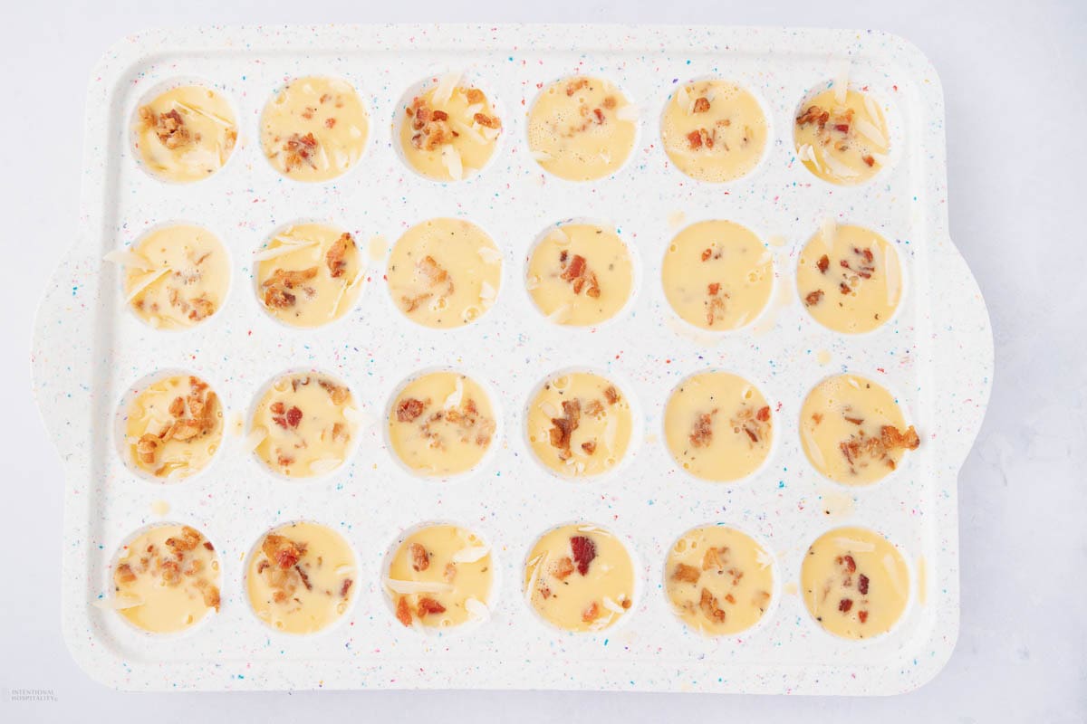 A white speckled baking tray with twenty-four circular compartments is filled with a creamy yellow mixture, topped with bits of bacon and grated cheese.