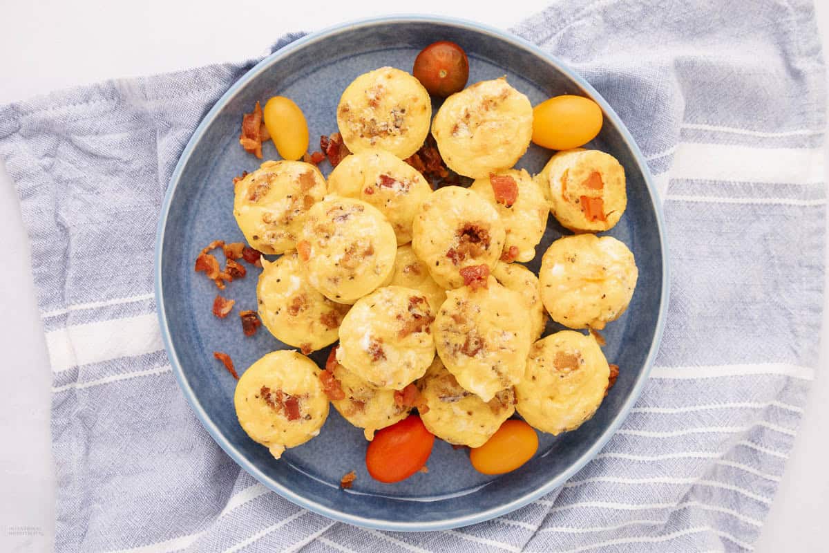 A plate of small savory muffins topped with bits of bacon, placed on a blue and white striped cloth. Several small yellow and red grape tomatoes are scattered around the muffins.