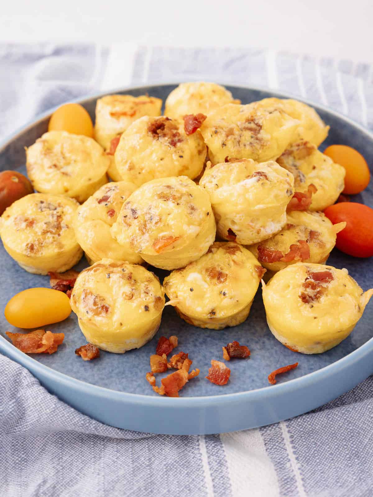 A blue plate filled with mini egg muffins, topped with bacon bits, surrounded by small orange and red tomatoes. The plate is set on a light blue, striped cloth.