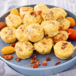 A blue plate piled with mini egg muffins featuring visible crumbles of bacon and melted cheese. Small cherry tomatoes in yellow and red are scattered around the muffins, adding color to the dish. The background shows a blue and white cloth.