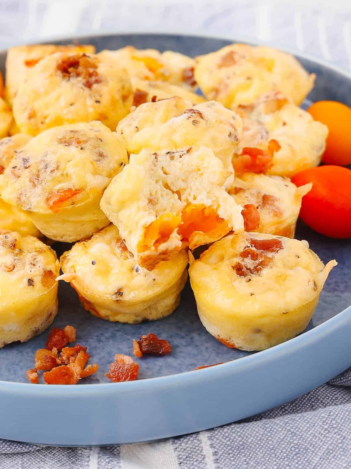 A blue plate filled with several small breakfast muffins, featuring bits of bacon and cheese. One muffin is split open, revealing a soft, fluffy interior. Two small red and orange tomatoes are on the plate as garnish.