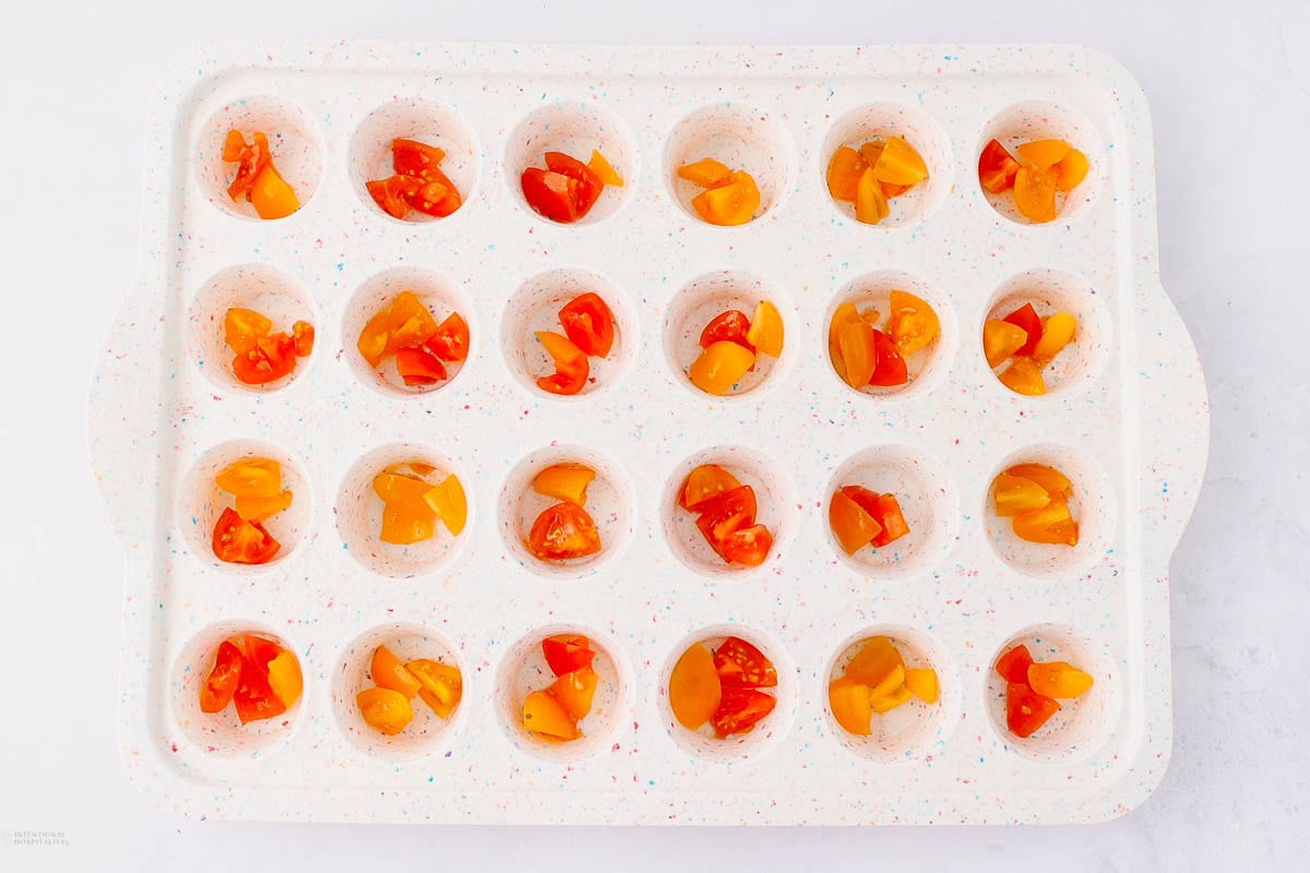 A white ice cube tray filled with diced red and orange cherry tomatoes in each compartment, placed on a white surface.