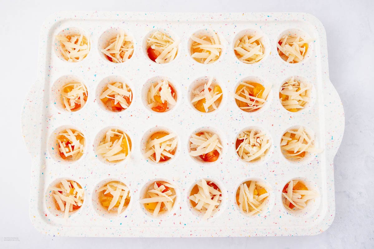 A white speckled ceramic tray holds 24 small, round compartments filled with eggs and topped with shredded cheese, ready for baking. The compartments are evenly arranged in four rows of six.