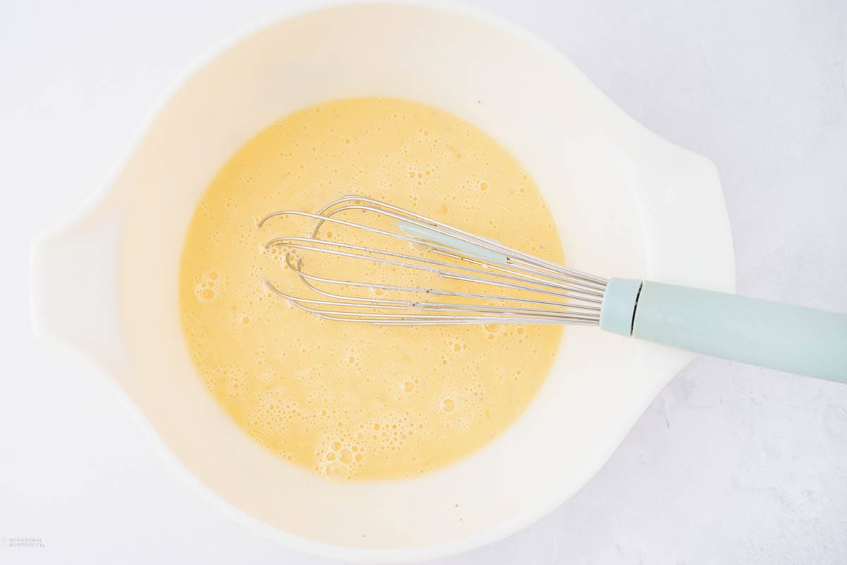 A white mixing bowl filled with whisked eggs, with a metal whisk featuring a light blue handle resting inside. The background is a light, neutral surface.