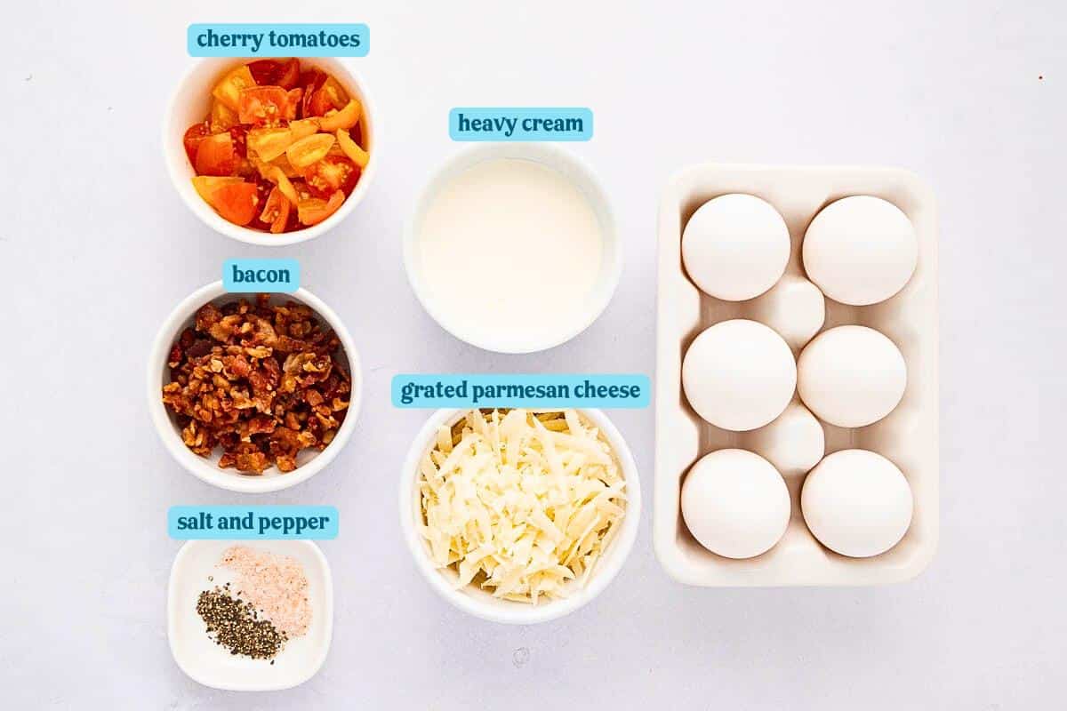 Ingredients on a white surface: a bowl of chopped cherry tomatoes, a bowl of crispy bacon bits, a small cup of heavy cream, a bowl of grated Parmesan cheese, a dish of salt and pepper, and a tray with six white eggs.