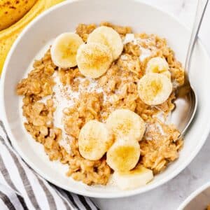 A bowl of oatmeal topped with banana slices and a sprinkle of cinnamon. A spoon is resting on the side of the bowl, and a banana and striped cloth are visible in the background.