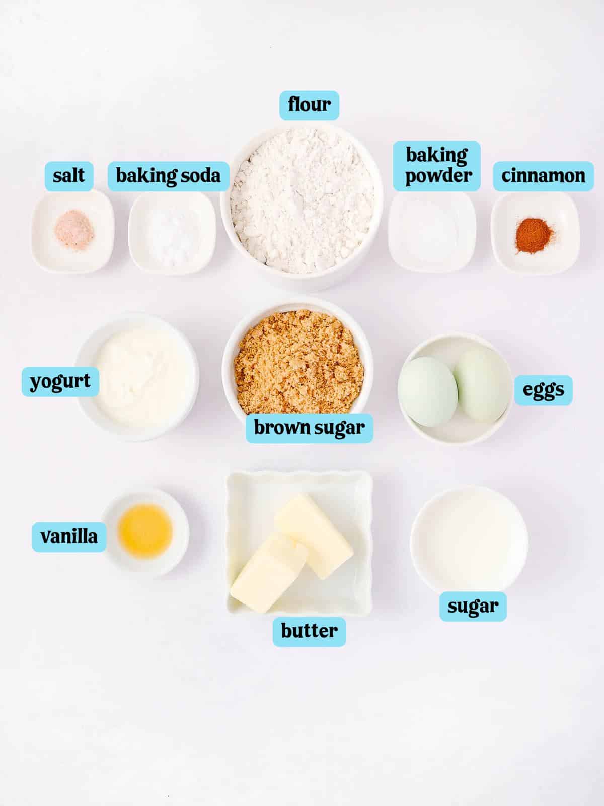 A collection of baking ingredients on a white surface, includes labeled containers of flour, baking soda, baking powder, cinnamon, salt, yogurt, brown sugar, eggs, vanilla, butter, and sugar.