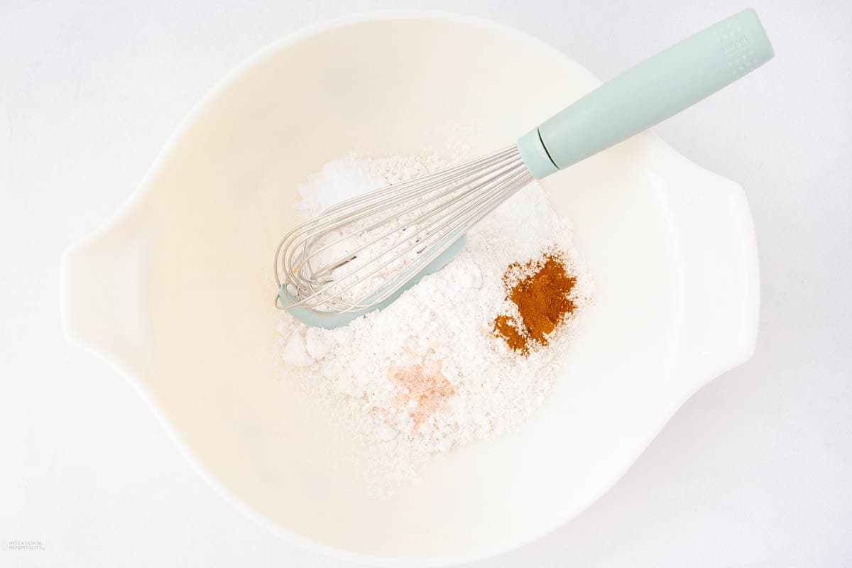 A mixing bowl containing flour, baking powder, cinnamon, and a pinch of salt, with a whisk resting on top.