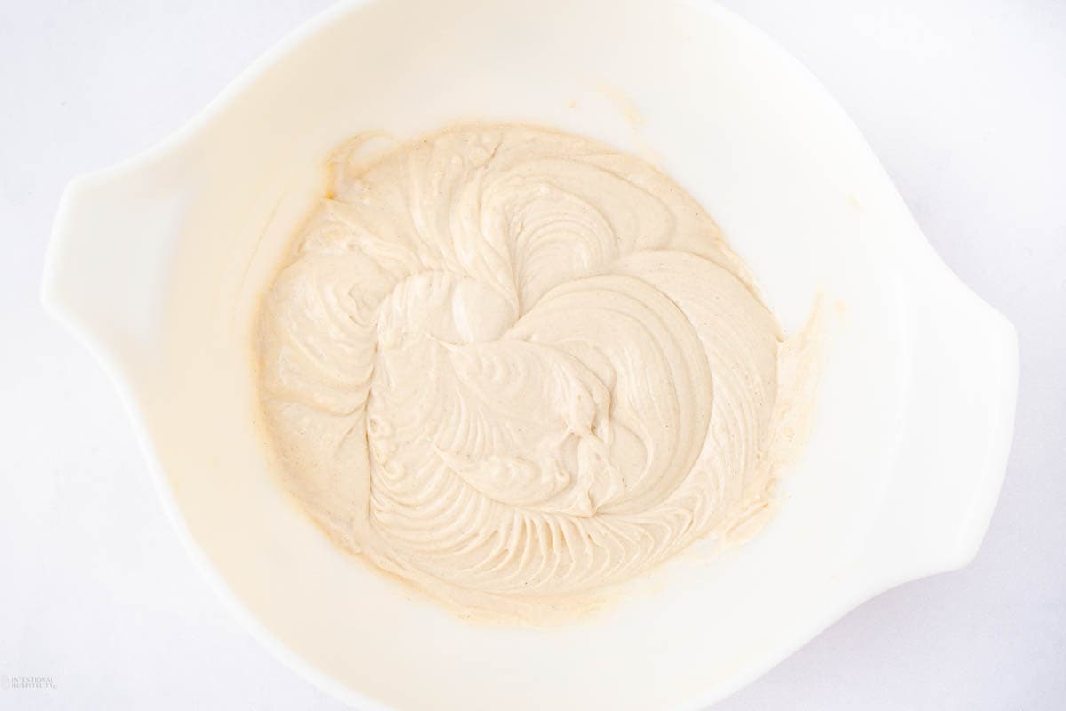 Creamy batter in a white mixing bowl, swirled smoothly with a light beige color, on a white surface.