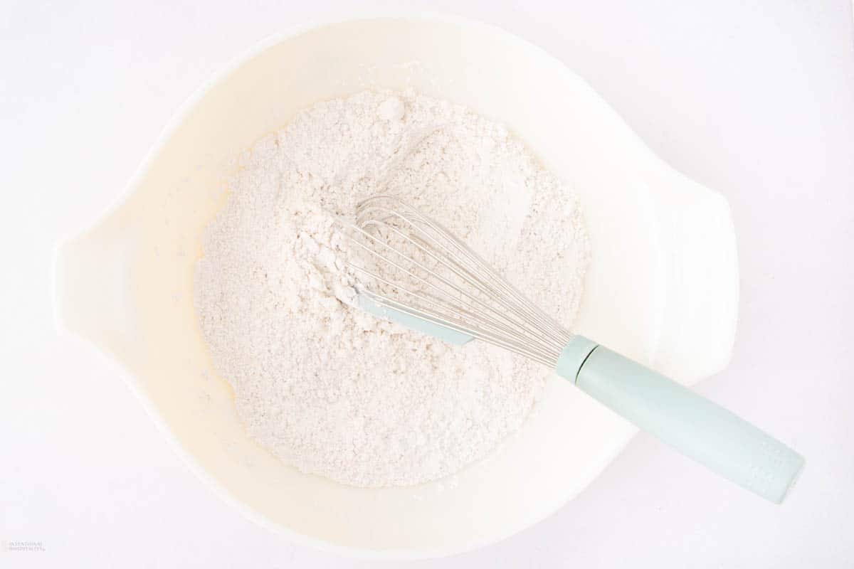 A whisk rests on top of a pile of flour in a large, off-white mixing bowl. The bowl is positioned on a white surface, and the flour appears finely milled, creating a smooth, soft texture.