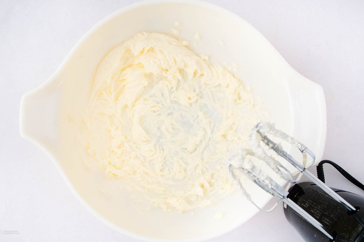 A white mixing bowl containing fluffy, creamed butter and sugar being mixed with an electric hand mixer. The mixer has two beaters that are partially covered with the mixture.