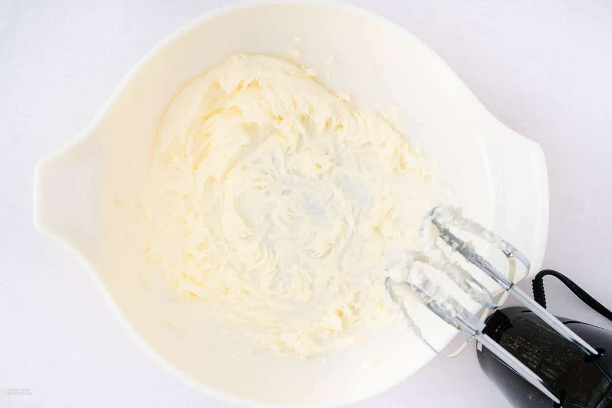 An electric mixer is blending butter and cream cheese in a white mixing bowl. The creamy mixture is swirled in the center of the bowl.