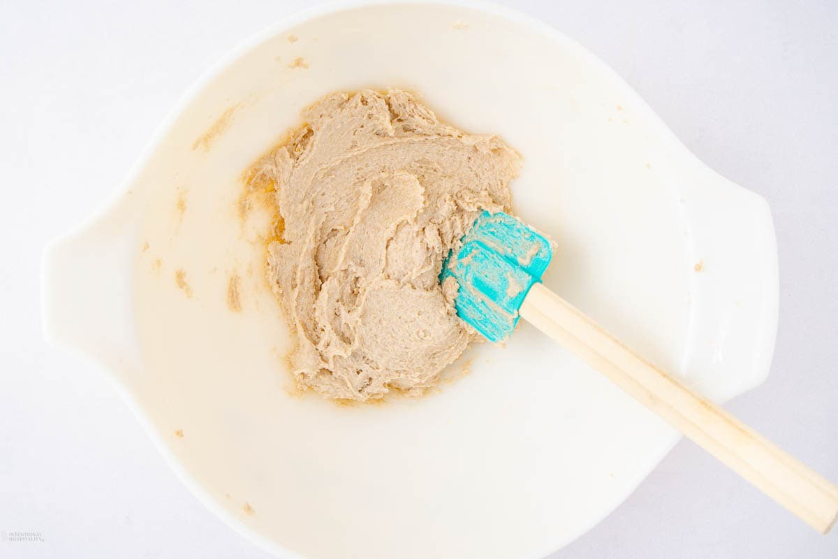 A white mixing bowl containing light brown dough. A turquoise rubber spatula with a wooden handle is resting in the bowl. The bowl is set against a simple, white background.