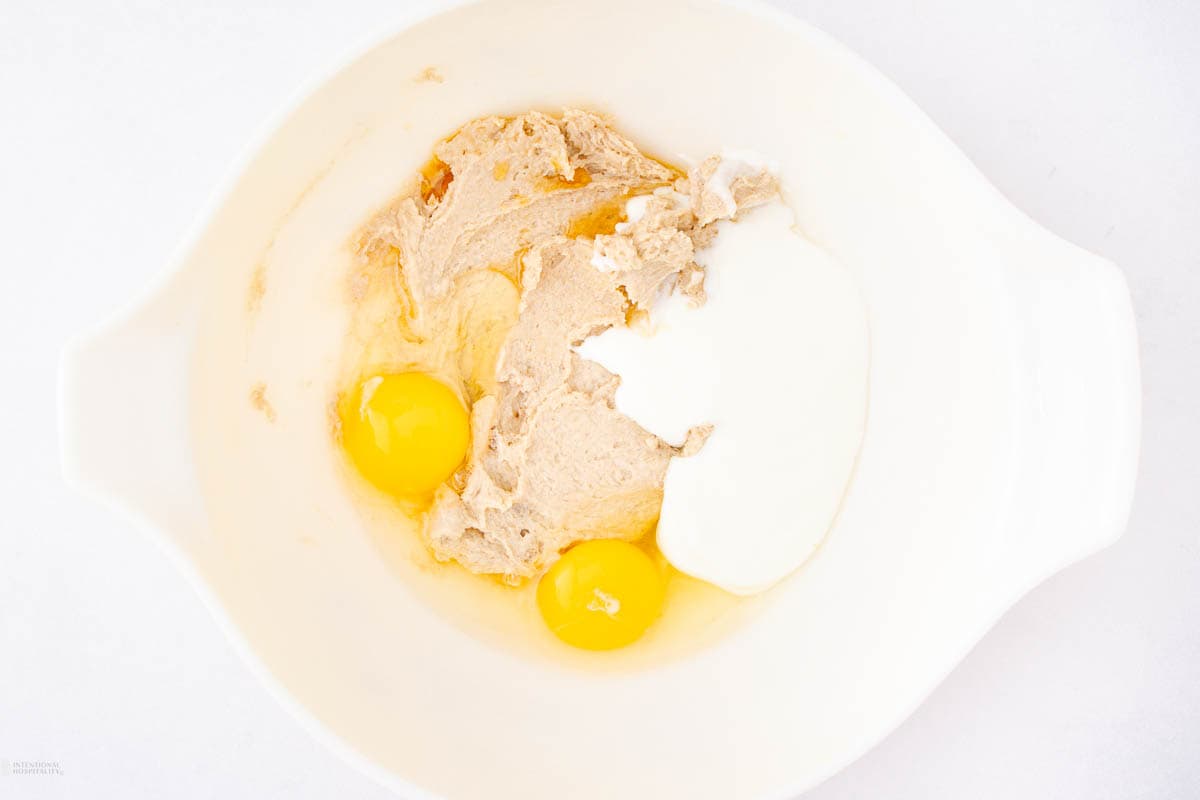 A white bowl containing two cracked eggs, brown sugar, and sour cream, ready to be mixed. The ingredients are partially combined, showing a blend of wet and dry textures on a light background.